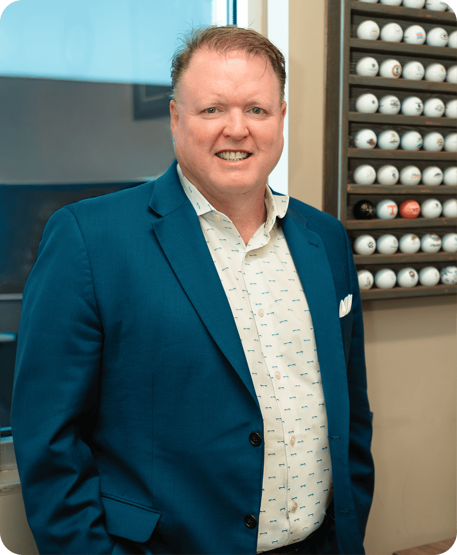 Dr. Kurt Steele smiling in a blue suit jacket and patterned shirt, standing in front of a display of golf balls.